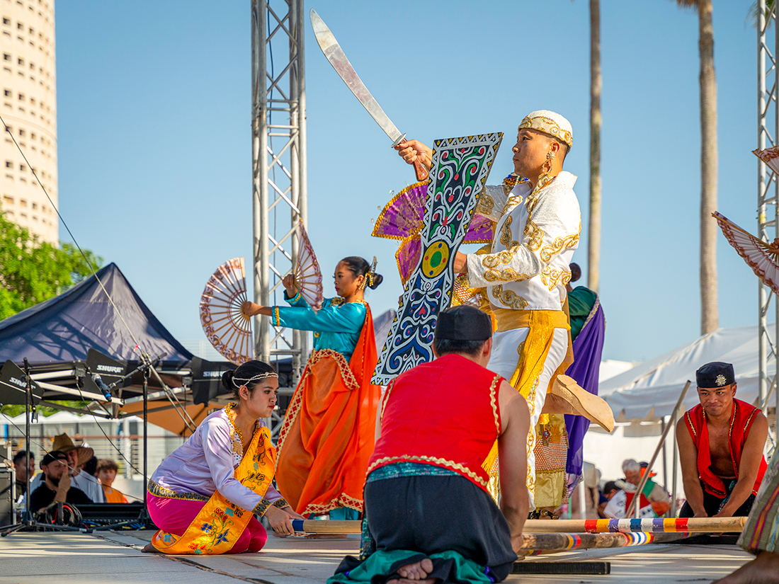 Male Dancer holding shield and pointing sword into the air.Female dancer with two fans. Several other dancers kneeling on ground holding long bamboo logs.