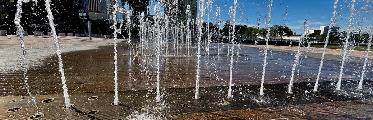 Jets of water shoot from a ground fountain in a city plaza, with tall buildings in the background.
